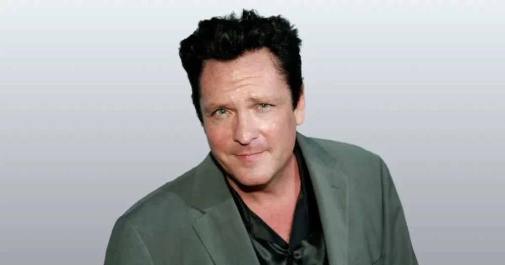 Michael Madsen death - A headshot of actor a man with dark, slightly disheveled hair and a subtle smirk, wearing a dark shirt under a grey blazer, against a plain grey background.