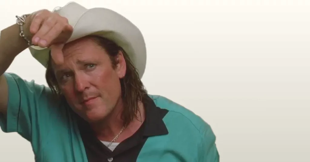 Michael Madsen - Cowboy Hat and Casual Look A close-up of actor Michael Madsen wearing a white cowboy hat, a turquoise shirt over a dark t-shirt, and a chain necklace. He looks directly at the camera with a subtle expression, while raising his right hand near his forehead.