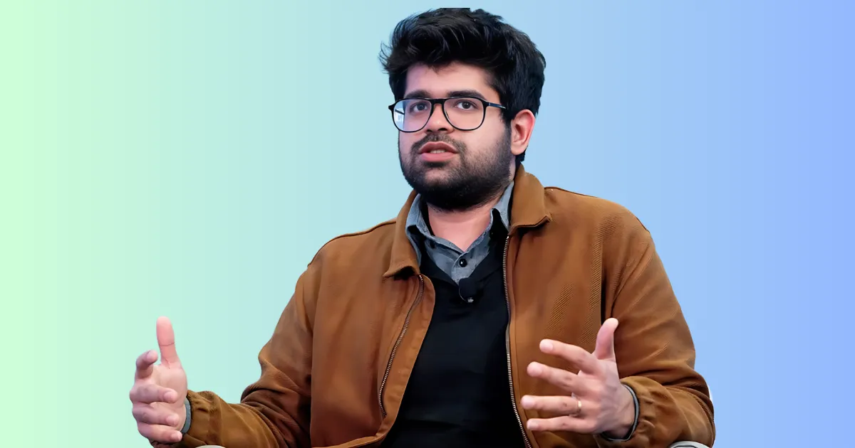 Aravind Srinivas, CEO of Perplexity AI, a young man with dark hair and glasses, wearing a brown jacket over a dark shirt, gestures with his hands while speaking, on a gradient blue-green background.