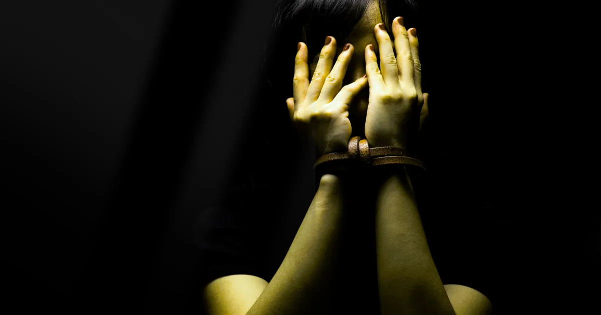 Los Angeles Fake ICE Kidnapping Scandal: A person's face is obscured by their hands, which are bound at the wrists with a brown strap, against a dark, shadowy background, conveying a sense of fear, captivity, or shame.